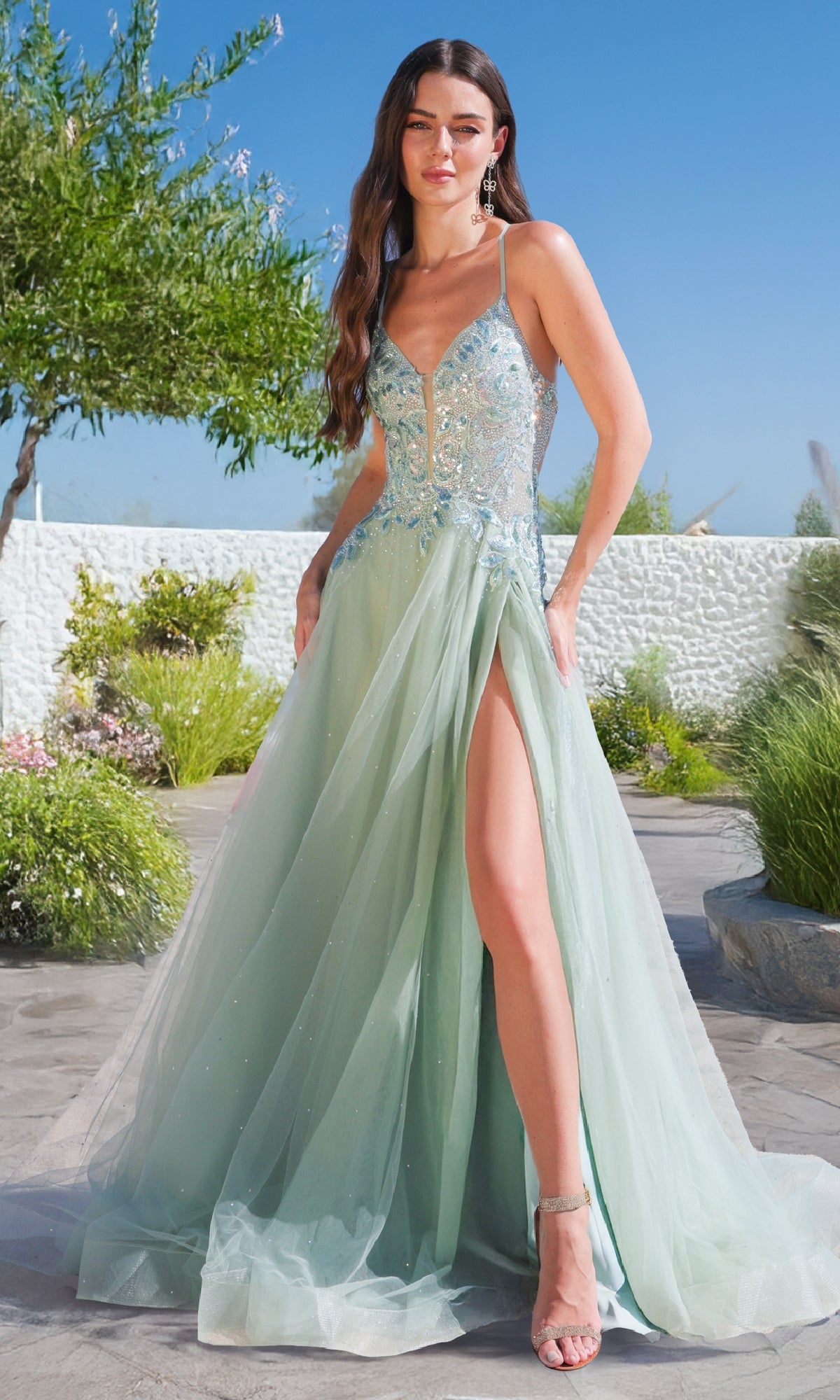 A woman stands outdoors among greenery and white stone walls, wearing the Amelia Couture TM1028 Long A-Line Prom Dress in light blue with embellishments, a sleeveless design, and a high slit.